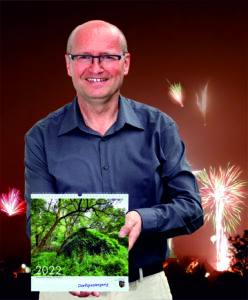 Ortsbürgermeister Matthias Mahl präsentiert den Heimatkalender 2022 vor dem Motiv des Monats Januar, dem Silversterfeuerwerk über Hütschenhausen.
